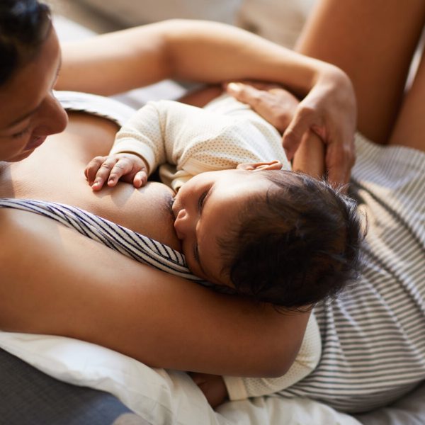 Close-up of serene loving young black mother in nightgown sitting on bed and breastfeeding baby in comfortable position