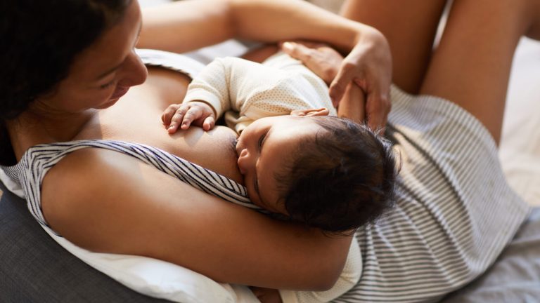 Close-up of serene loving young black mother in nightgown sitting on bed and breastfeeding baby in comfortable position