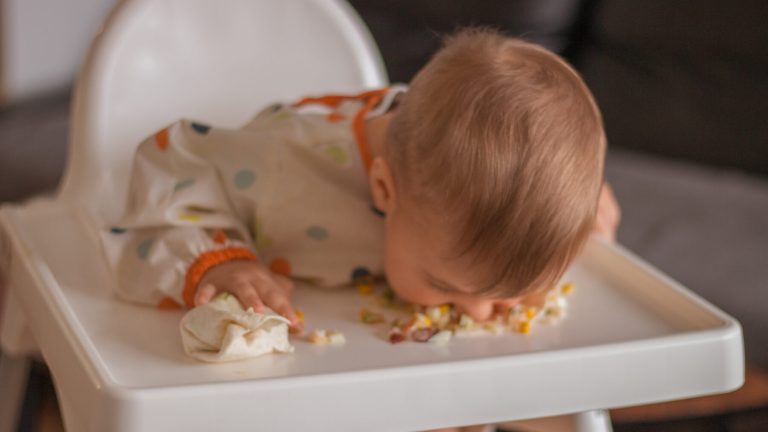 Baby boy sitting in highchair and eating healthy lunch with his head bent. Baby learning to eat using his mouth.