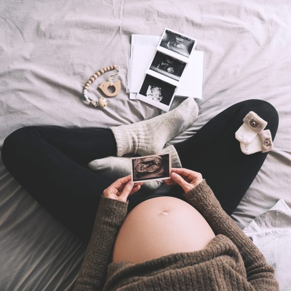 Pregnant woman holding ultrasound image looking at medical test reports in cozy knitted sweater in bed at cold winter days. Expectant mother with pregnant belly waiting for baby. Concept of pregnancy.